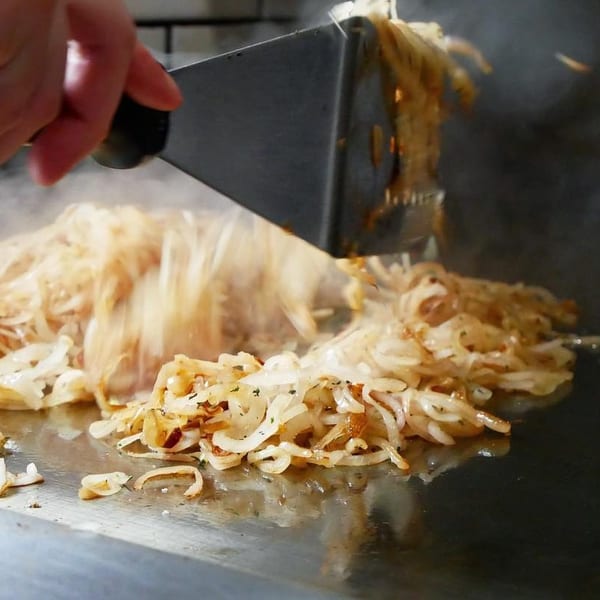 A hand uses a spatula to flip sizzling, caramelized onions on a flat-top grill.