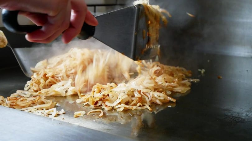 A hand uses a spatula to flip sizzling, caramelized onions on a flat-top grill.