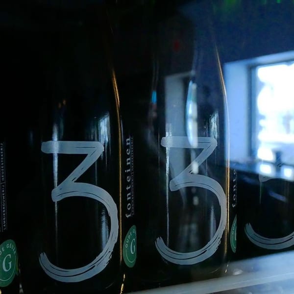 Row of dark beer bottles with "3 Fonteinen" labels, reflecting a bright window and blurred person.
