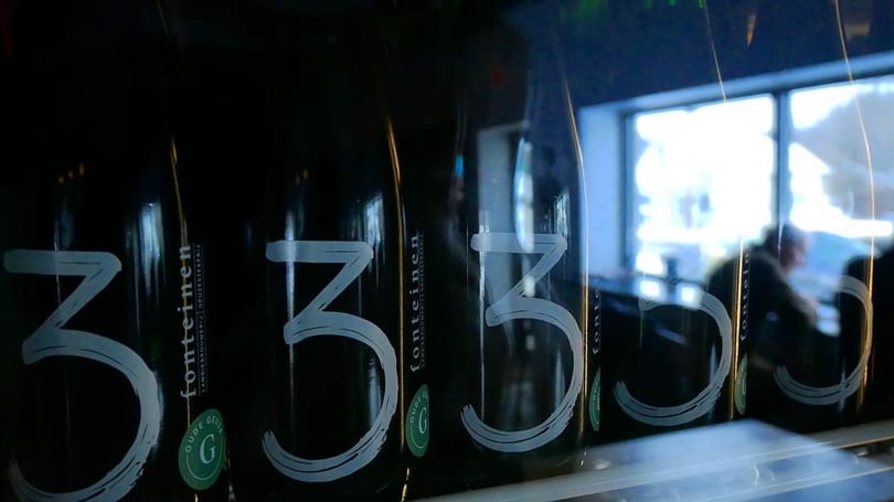Row of dark beer bottles with "3 Fonteinen" labels, reflecting a bright window and blurred person.