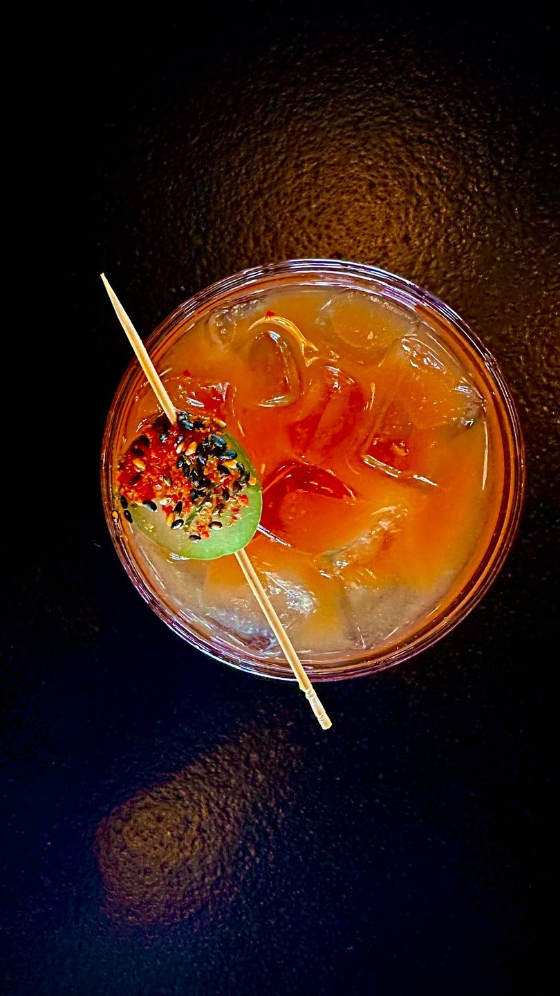 Overhead view of a vibrant orange cocktail with ice, garnished with a spiced cucumber slice.