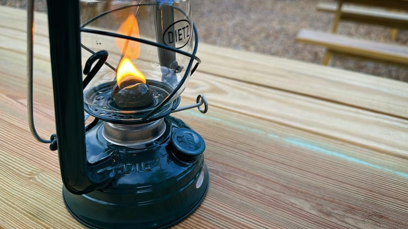 A lit green Dietz lantern sits on a wooden picnic table, providing warm light.