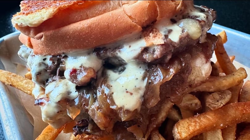 A close-up of a cheesesteak sandwich with melted cheese and caramelized onions, served with fries.