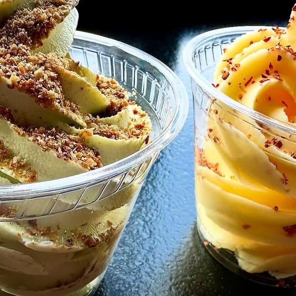 Two cups of soft-serve ice cream, one green with nuts, one yellow with red flakes.