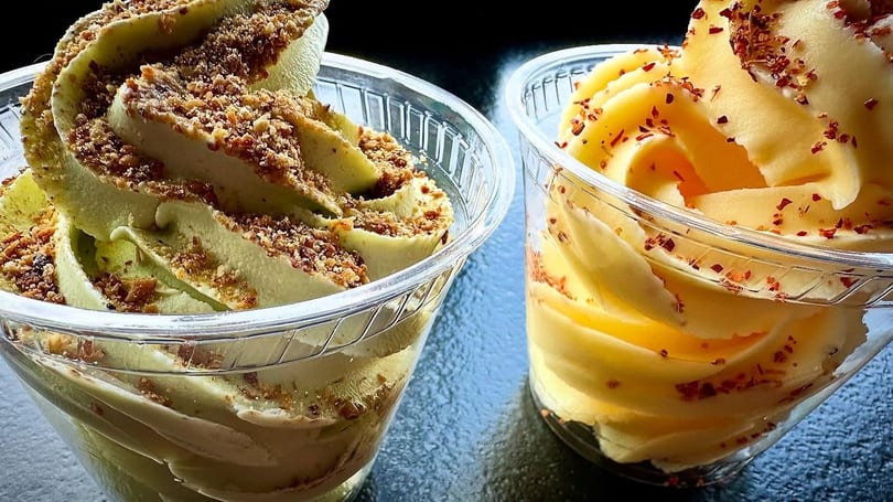 Two cups of soft-serve ice cream, one green with nuts, one yellow with red flakes.