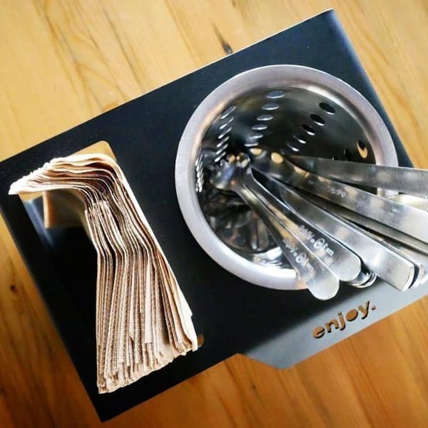 Black caddy with brown napkins and a metal holder filled with silverware on a wooden table.