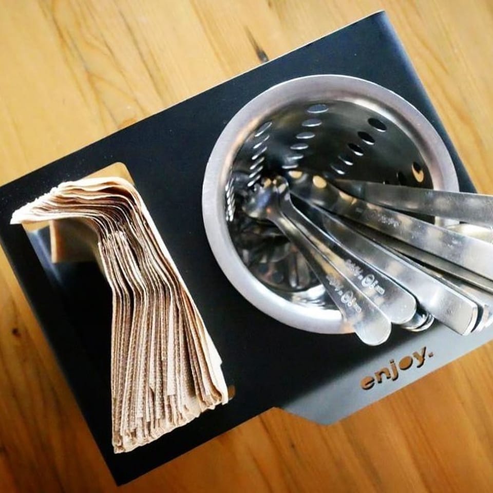Black caddy with brown napkins and a metal holder filled with silverware on a wooden table.