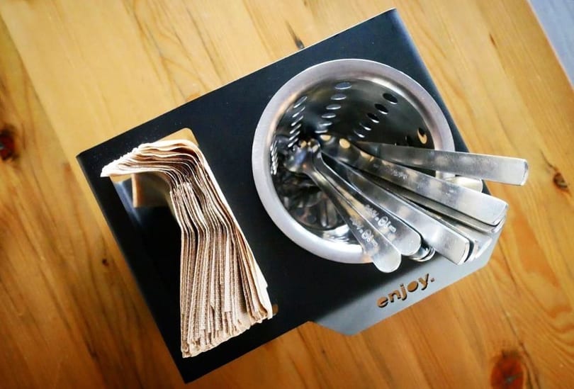 Black caddy with brown napkins and a metal holder filled with silverware on a wooden table.