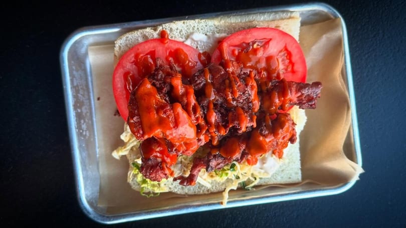 Spicy fried chicken sandwich with tomatoes, lettuce, and sauce on a metal tray.