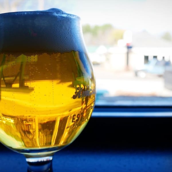 A glass of golden beer with foamy head sits on a dark surface, with a blurred outdoor background.