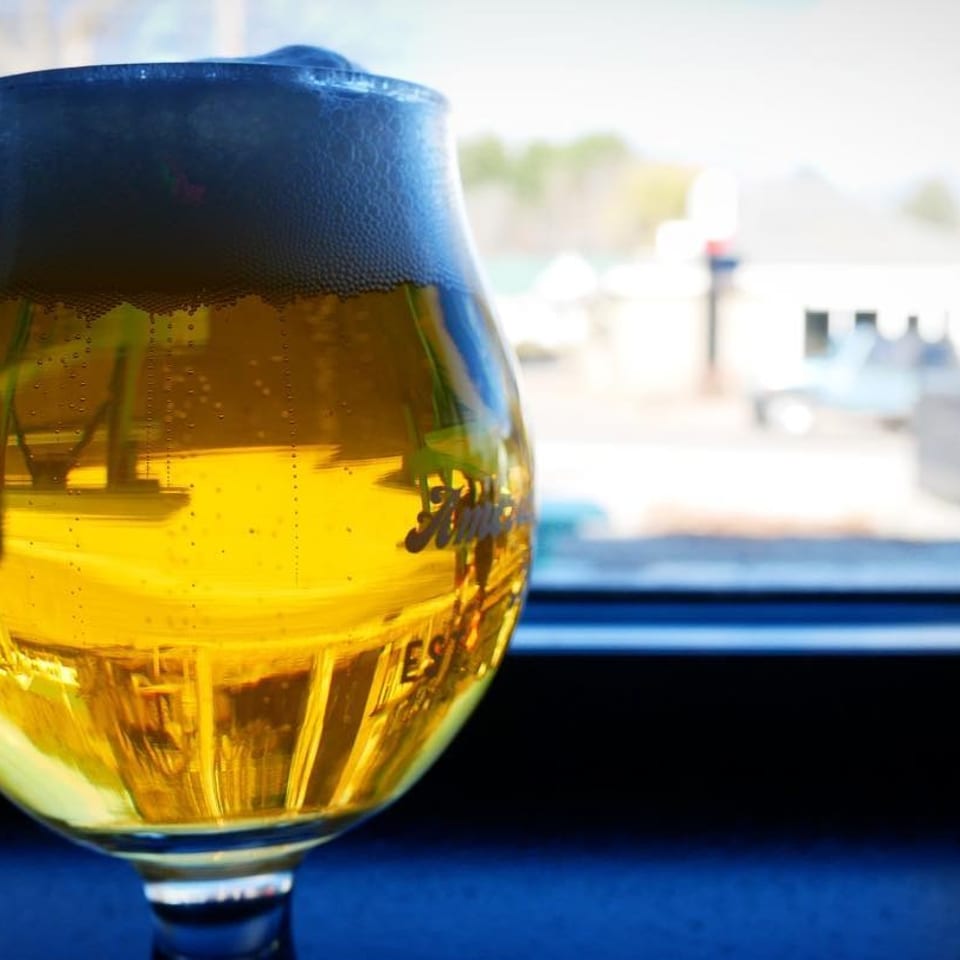 A glass of golden beer with foamy head sits on a dark surface, with a blurred outdoor background.