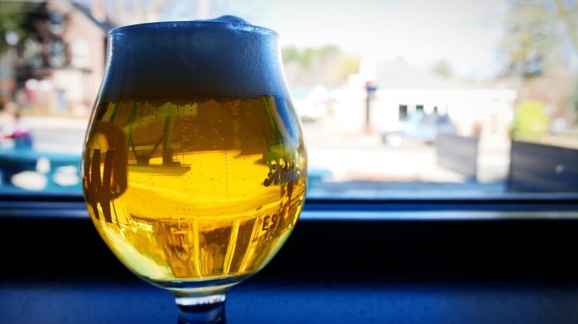 A glass of golden beer with foamy head sits on a dark surface, with a blurred outdoor background.